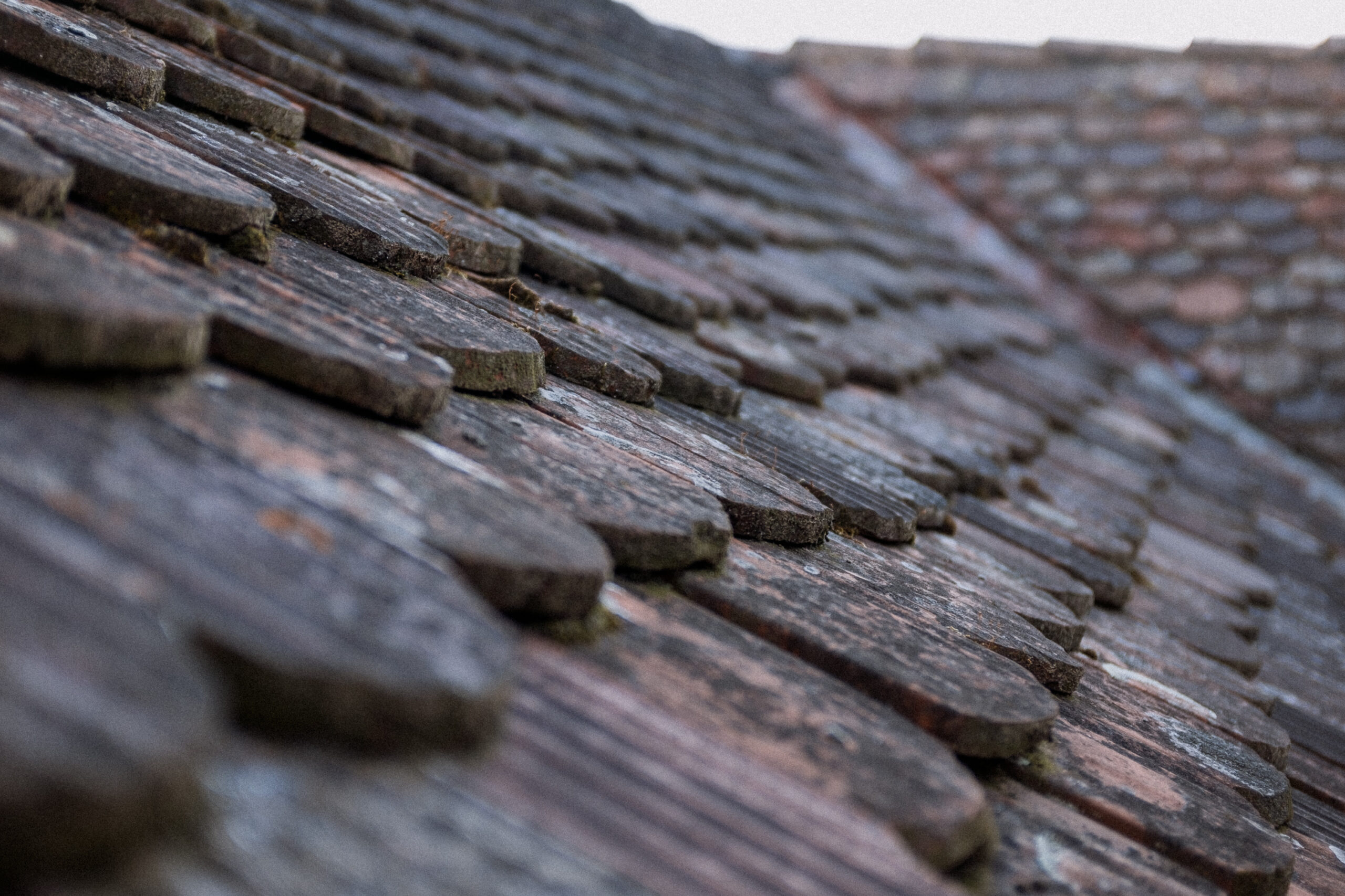 This image shows a close-up view of an aged and weathered roof made of wooden shingles. The shingles have a natural, rustic appearance with visible wear, moss growth, and a textured surface. The angle highlights the overlapping pattern of the shingles, emphasizing their layered structure.