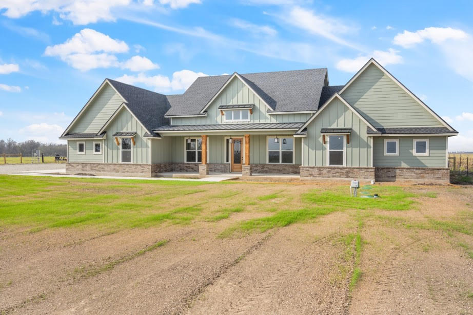 This image displays a modern single-story house with a green exterior and a sloped asphalt-shingle roof, similar to the one in the previous image. The house has a combination of board-and-batten siding and stone accents along the bottom. The front features a prominent entrance with wooden columns and large windows. It is set on a spacious lot with partially grown grass and a blue sky overhead. 